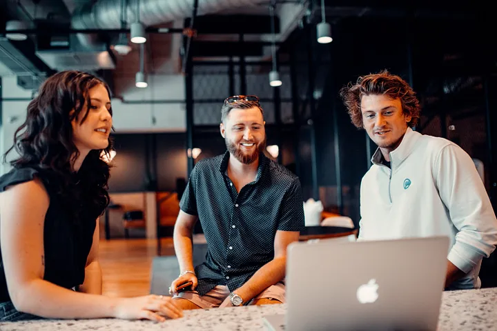 Three smiling business people sit in an industrial setting, appearing to verbally interact with a laptop.
