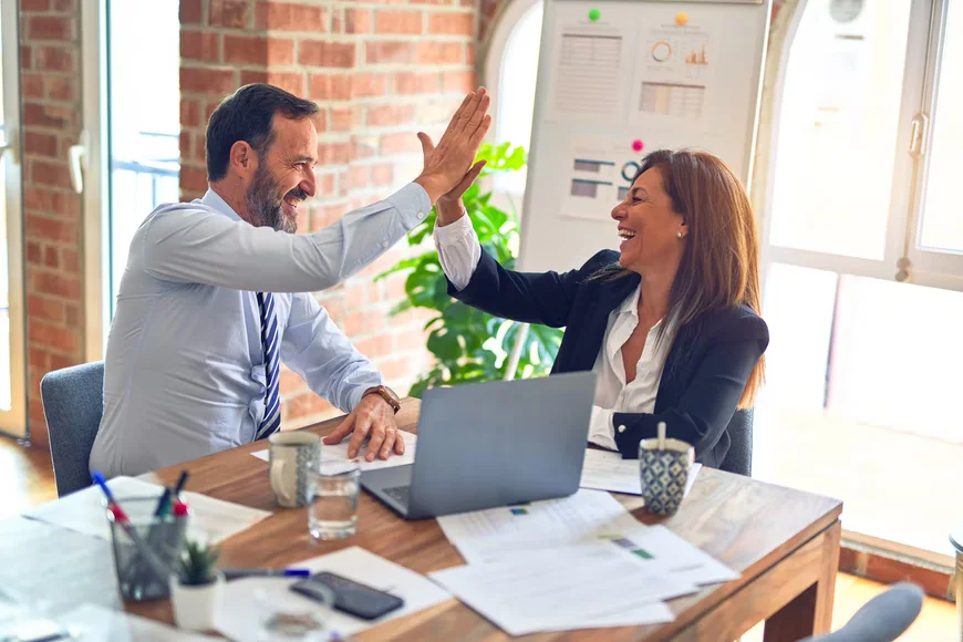 Two people are high fiveing each other. They are wearing business suits and sit at a conference table covered with coffee cups, paperwork, laptop, calculator, and other office accoutrement. In the background is an easel holding graphs.